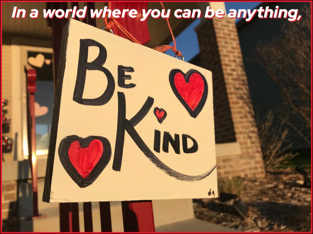 In a world where you can be anything, be kind. A photo of a painted sign by Doris Gassen that says Be Kind with red hearts on it. 